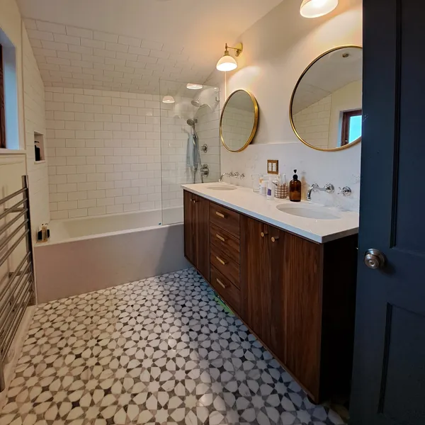 Bathroom with a walnut double vanity, round brass mirrors, subway-tile tub surround and a marble mosaic floor